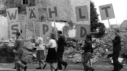 Vor den Trümmern eines Hauses tragen junge Männer und Frauen Schilder, die den Schriftzug "Wählt SPD" ergeben, Juni 1946