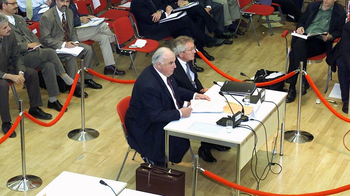 Helmut Kohl sitzt neben einem anderen Mann an einem Tisch. Der Bereich um den Tisch ist mit roten Seilen abgesperrt, dahinter sitzen Fotografen und Journalisten.