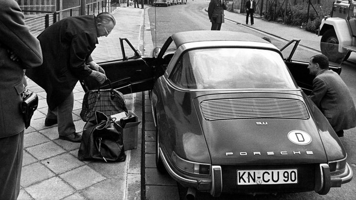 Polizisten schauen zu, wie zwei Männer einen dunklen Porsche 911 durchsuchen. Die Türen sind offen, auf dem Bürgersteig stehen mehrere Taschen.