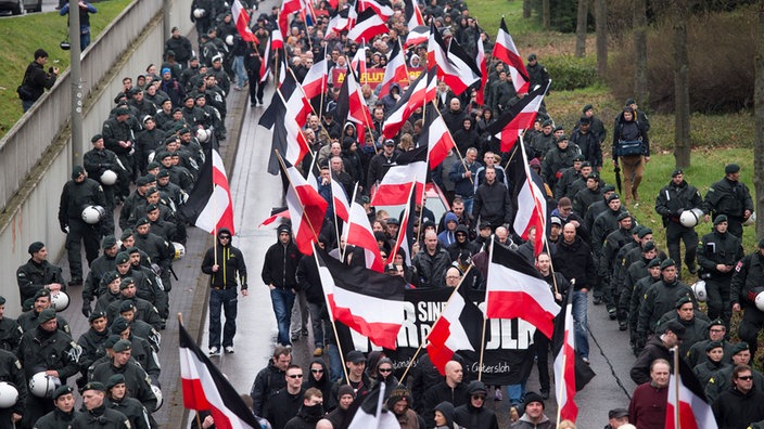 Demonstrationszug mit schwarz-weiß-roten Fahnen, flankiert von Polizeibeamten.