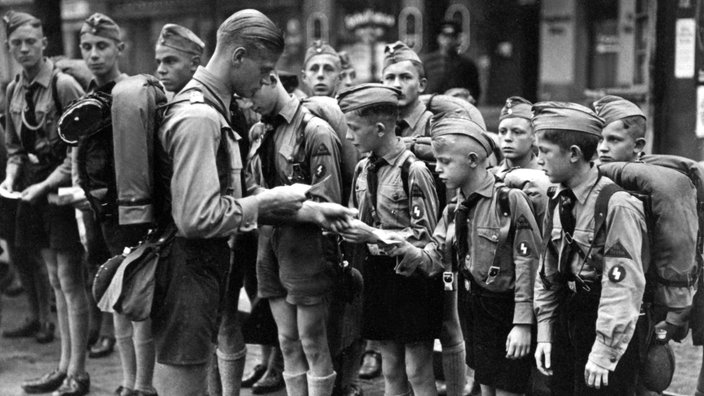 Eine Gruppe der 'Berliner Pimpfe' in Uniformen steht am Straßenrand und wird von einem Betreuer gemustert.