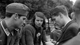 Sophie Scholl sitzt, Christoph Probst steht ihr gegenüber und notiert etwas auf einem Block. Beide schauen ernst. Im Hintergrund sind Bäume zu sehen.