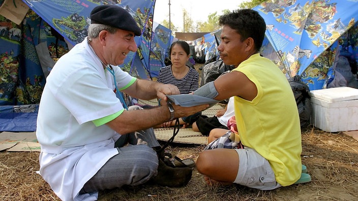 Ein italienischer Krankenpfleger sitzt zusammen mit einem thailändischen Mann auf dem Boden und misst diesem den Blutdruck.