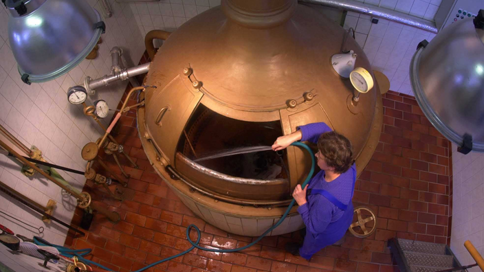 Eine Frau spritzt mit einem Schlauch Wasser in einen großen, kupferfarbenen Kessel.