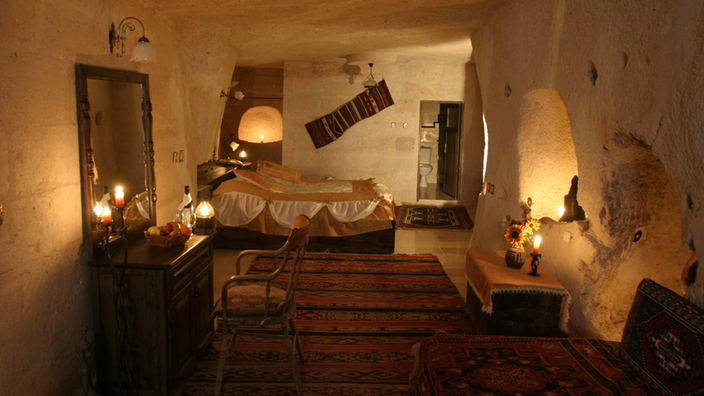 Hotelzimmer in einer Höhle in Kappadokien. Die Wände sind weiß gekalkt und schief. Überall sind kleine Lampen eingelassen. An den Wänden und auf dem Boden sind Teppiche zu sehen. Der Blick geht auf das Bett im Hintergrund und einen alten Schreibtisch mit Spiegel im Vordergrund links.