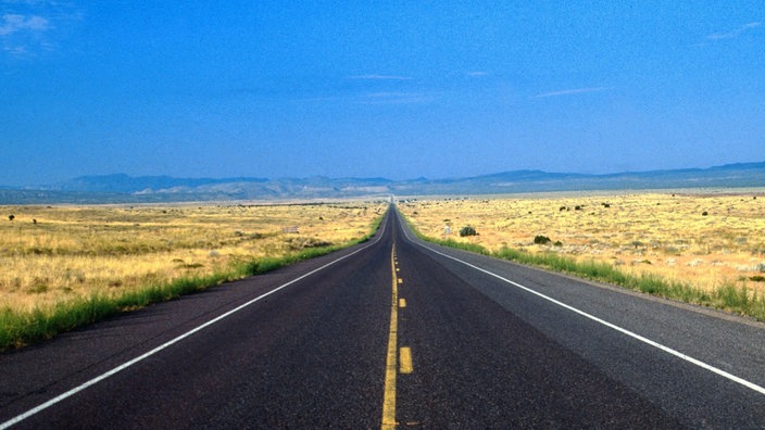 Eine schnurgerade Straße bis zum Horizont, rechts und links trockene Graslandschaft, im Hintergrund die Berge