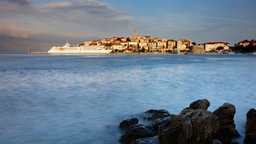 Blick übers Meer auf die historische Altstadt von Korčula, an der eine weiße Fähre vorbeifährt.