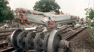 Die Achse eines Waggons liegt im Vordergrund des Bildes auf den Gleisen. Im Hintergrund ist die eingestürzte Brücke zu sehen. 