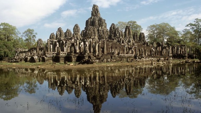 Das Bild zeigt einen großen Tempel mit vielen kleinen terrassenförmigen und unterschiedlich hohen Türmen. Er spiegelt sich in einem Wassergraben im Vordergrund.