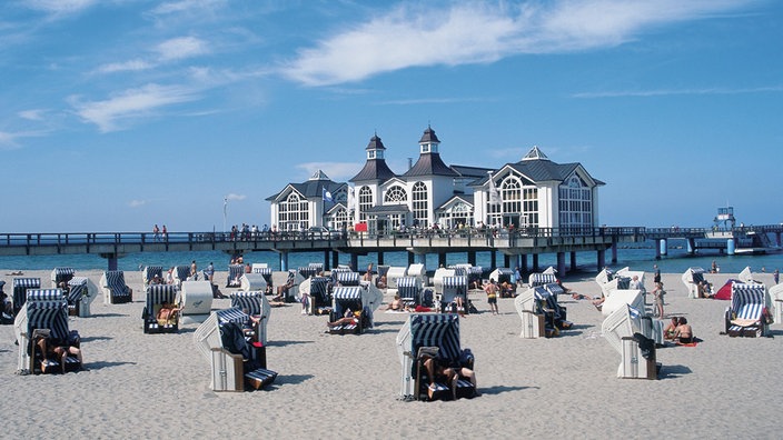 An einem Sandstrand sitzen Menschen in Strandkörben. Im Hintergrund steht ein weißes Haus mit roten Türmen auf Stelzen im Wasser.