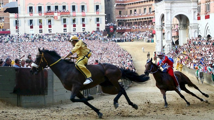 Zwei Rennpferde mit Jockeys in einer Kurve auf einem mittelalterlichen Platz. Im Hintergrund zahlreiche Besucher im Innenraum des Platzes und an den Fenstern und Balkonen der umliegenden Häuser.