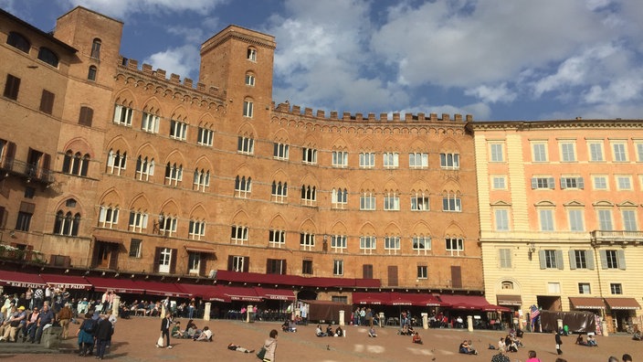 Blick auf den Platz 'Il Campo' in Siena während der Dämmerung. Auf dem großen Platz sind vereinzelt Menschen zu sehen; im Hintergrund die Silhouetten von beleuchteten Häusern und der Turm des Doms.