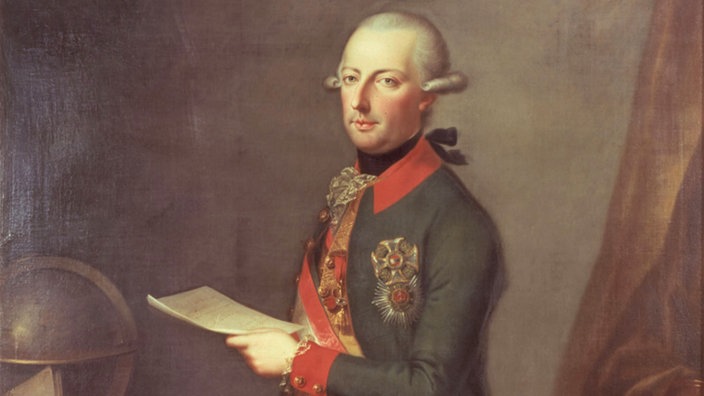 Kaiser Joseph II. in seiner kaiserlichen Tracht. Die Haare hat er der Mode nach zum Zopf gebunden. Er steht an seinem Schreibtisch, den ein Globus und eine Landkarte zieren.