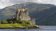 In einer wolkenverhangenen Landschaft thront das Schloss Eilean Donan Castle am Ufer eines Sees.