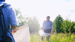 Zwei Menschen mit großen weißen Keschern stehen in einer Wiese mit hüfthohem Gras.