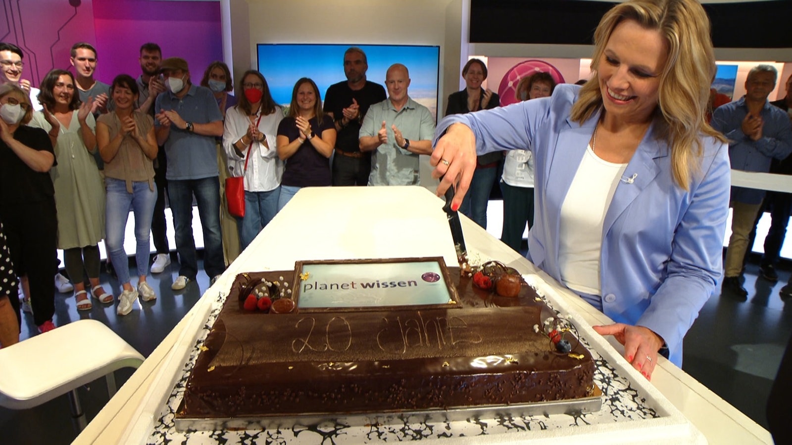 Planet-Wissen-Moderatorin Andrea Grießmann schneidet eine Torte an, dahinter stehen viele Menschen und applaudieren