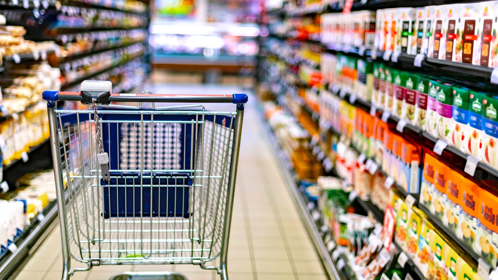 Fokus auf leeren Einkaufswagen zwischen vollen Supermarktregalen.