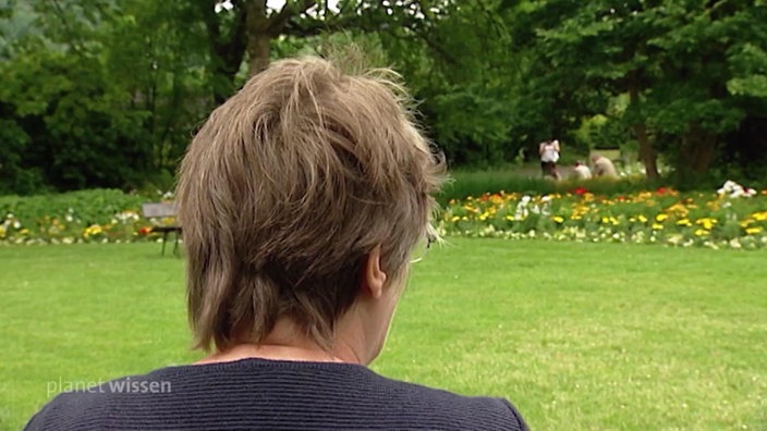 Eine Frau sitzt mit dem Rücken zur Kamera und blickt auf eine Wiese mit einem Blumenbeet.