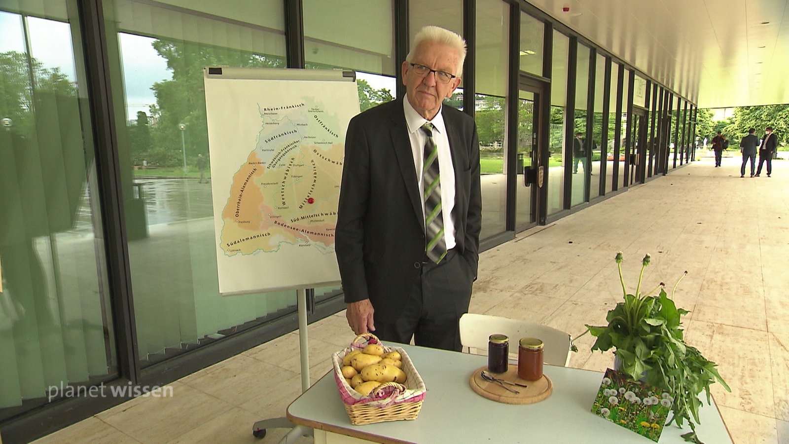 Der baden-württembergische Ministerpräsident Kretschmann steht vor einer Wandtafel mit einer Karte der Dialektregionen von Baden-Württemberg.