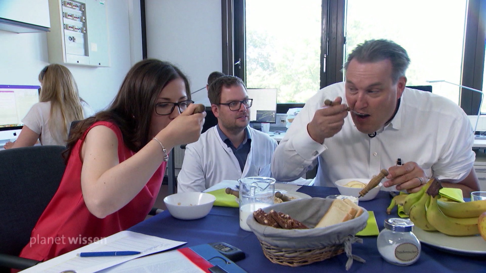 Eine Frau und ein Mann sitzen an einem Bürotisch und essen, während ein Mann in weißem Laborkittel ihnen zuschaut.