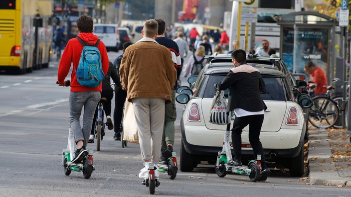 Eine Straße mit Bushaltestelle, vielen Parkenden Autos und Radfahren. E-Scooter-Fahrer setzen im Vordergrund zur Fahrt an